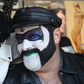Guard Inya from The San Diego Sisters of Perpetual Indulgence wearing clown white with blue and green glitter makeup, black leather cap, and black leather vest.